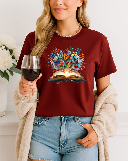 Person wearing a maroon t-shirt with a colorful book graphic, holding a glass of red wine.