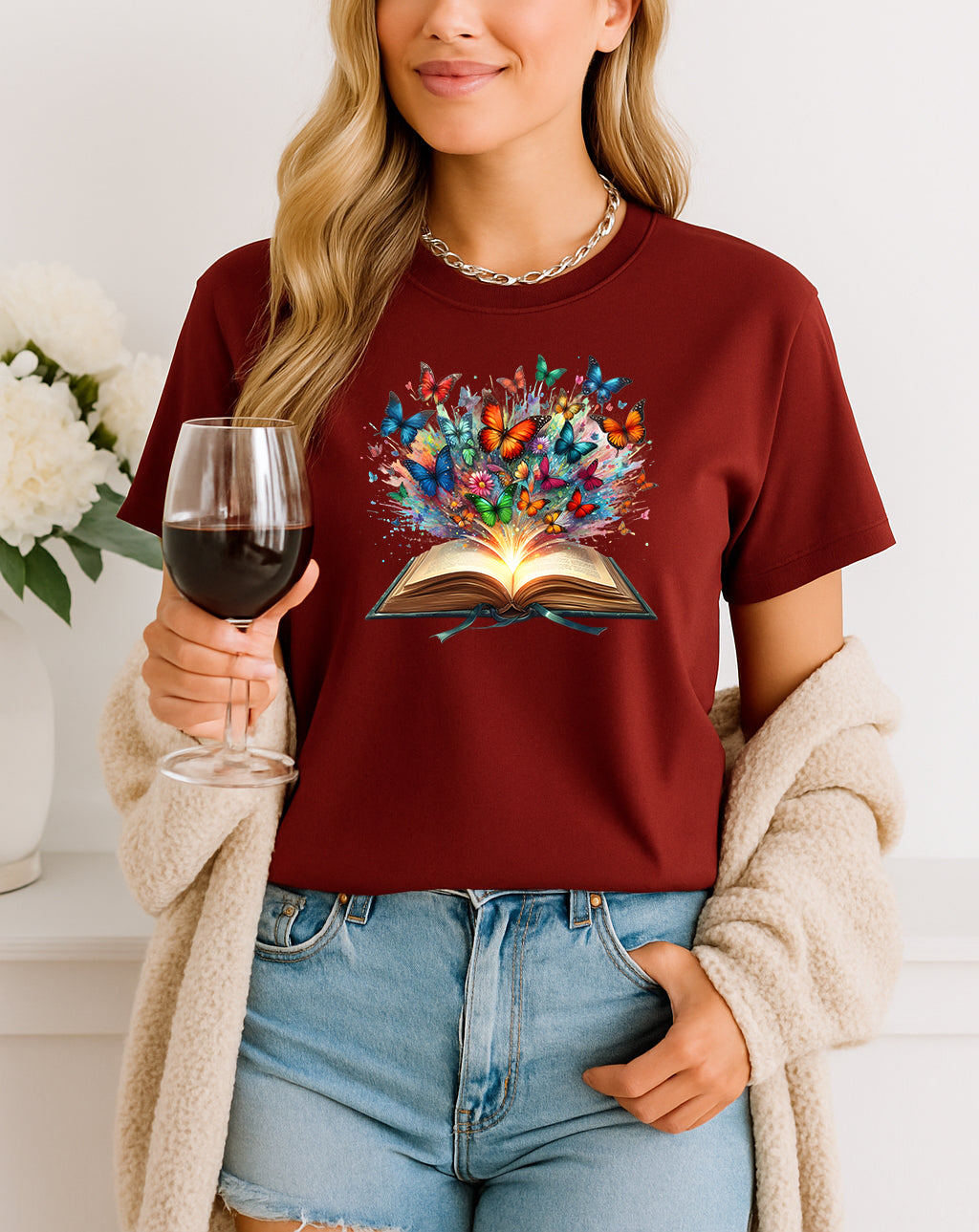 Person wearing a maroon t-shirt with a colorful book graphic, holding a glass of red wine.